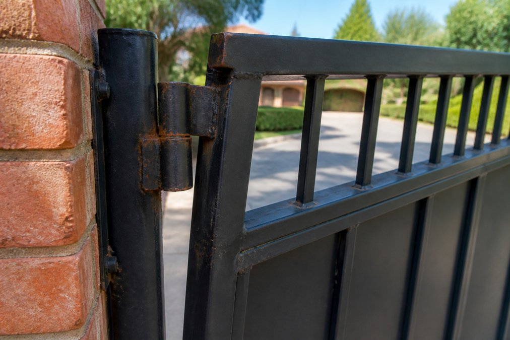 Close-up of a residential swing gate with visible rust on the hinges and a misaligned gate panel leaning away from the post, photographed in afternoon sunlight against a brick driveway pillar
