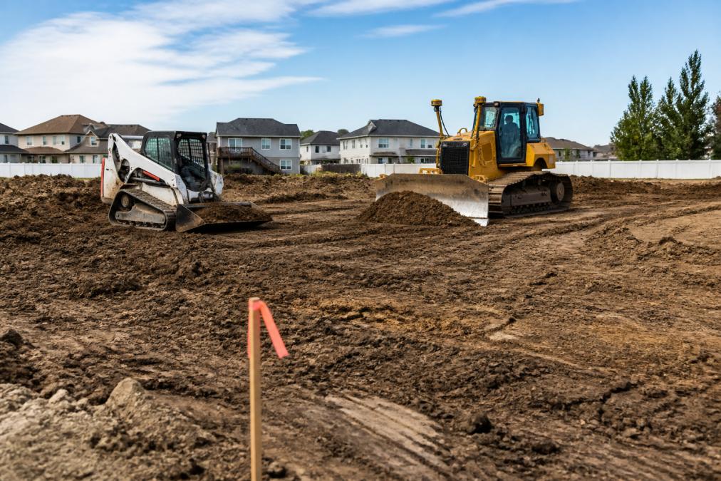 A skid steer and bulldozer working on a residential lot, moving topsoil and grading the subgrade for a new home foundation, with a site survey marker visible in the foreground