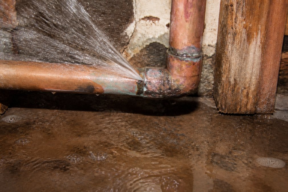 Close-up of a burst copper pipe with water actively spraying from the rupture point, water pooling on the floor beneath, showing visible corrosion and damage at the joint