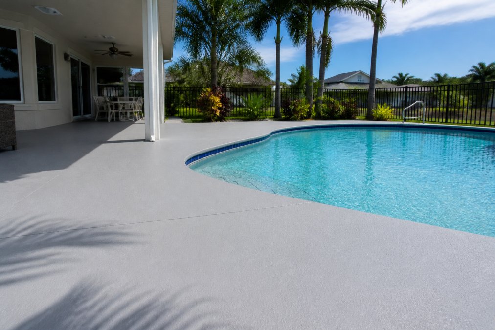A freshly resurfaced residential pool deck with a smooth slip-resistant acrylic coating in a light gray finish, surrounding a clean blue swimming pool on a bright Florida afternoon with palm trees visible in the background