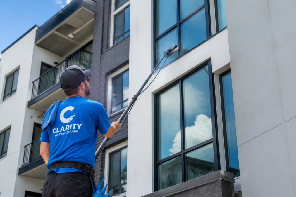 Professional window cleaner using a water-fed pole on the exterior of a multi-story residential condo building, residential balconies visible in the background under bright daylight