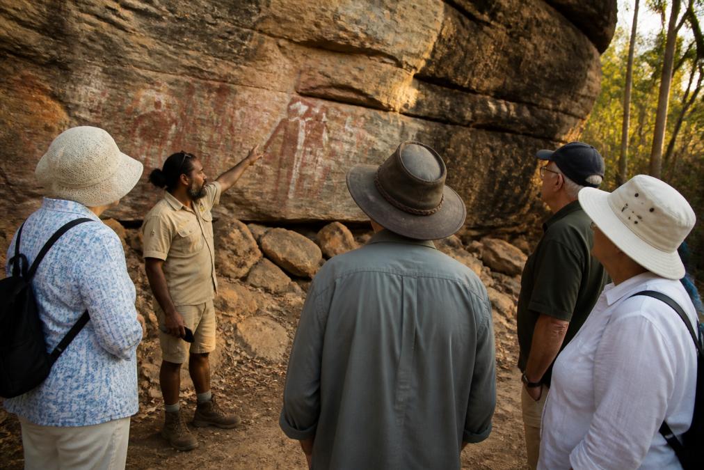 A small tour group of older travellers standing at a respectful distance listening to an Aboriginal guide who is pointing toward ancient rock art on a sandstone cliff in warm late-afternoon light