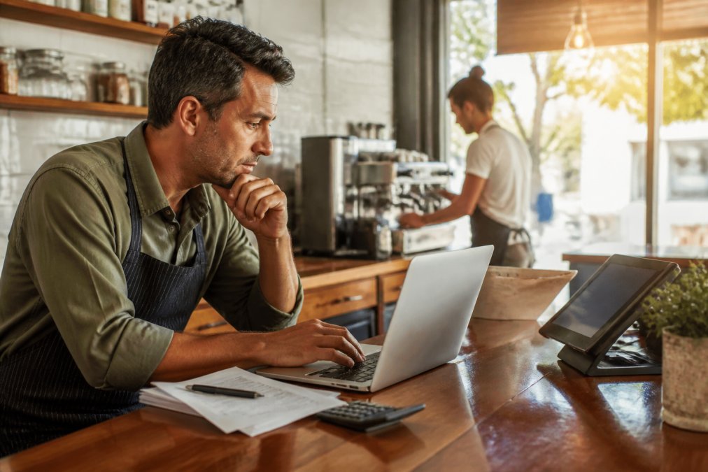 A cafe owner reviewing loyalty program pricing on a laptop at the counter, barista working in the background, tablet POS system visible on the counter, warm afternoon light through front windows