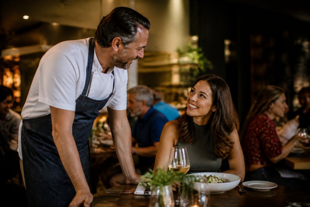 A restaurant owner speaking with a regular customer at their table, both smiling, with warm lighting and other diners visible in the soft-focused background