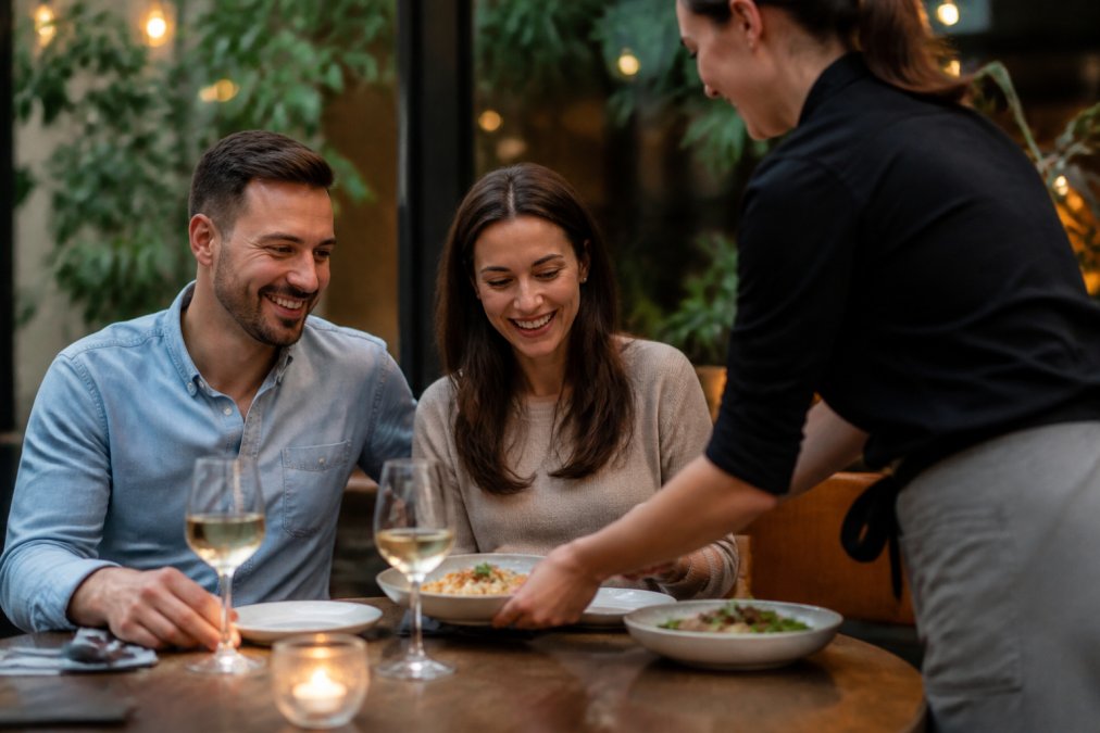 A server handing a warm, freshly prepared dish to a smiling couple at a cozy, well-lit neighborhood restaurant table