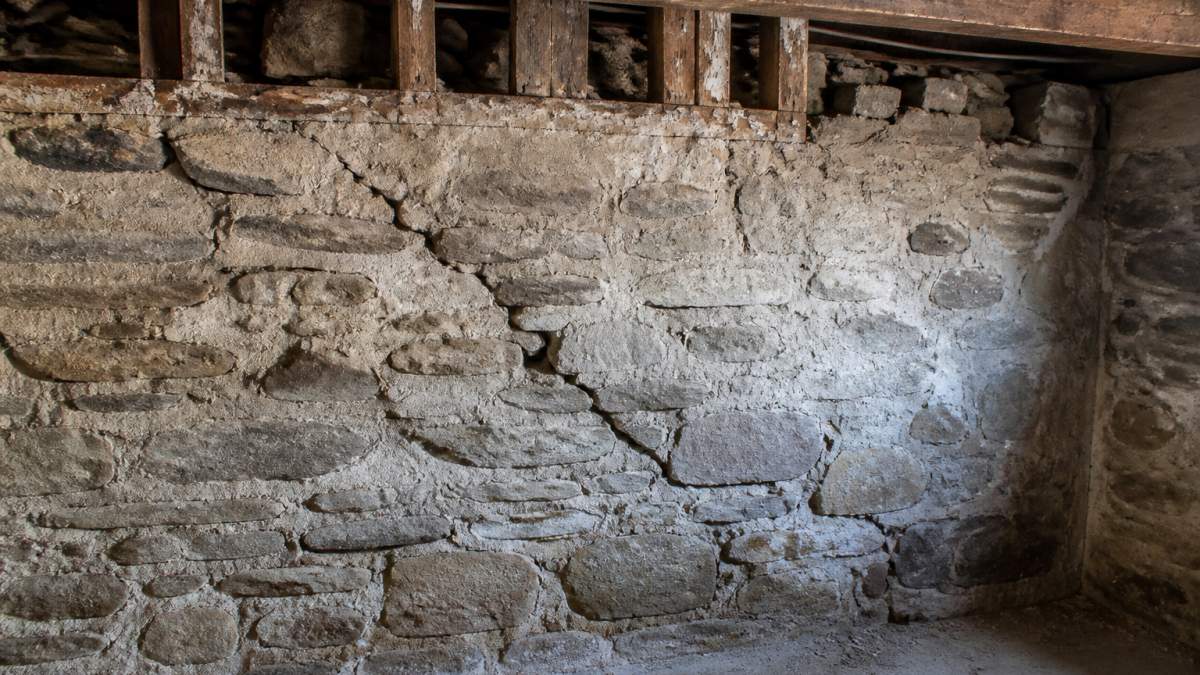 Close-up of cracked stone foundation wall showing diagonal settlement cracks and deteriorating mortar joints in a historic basement with exposed timber framing above
