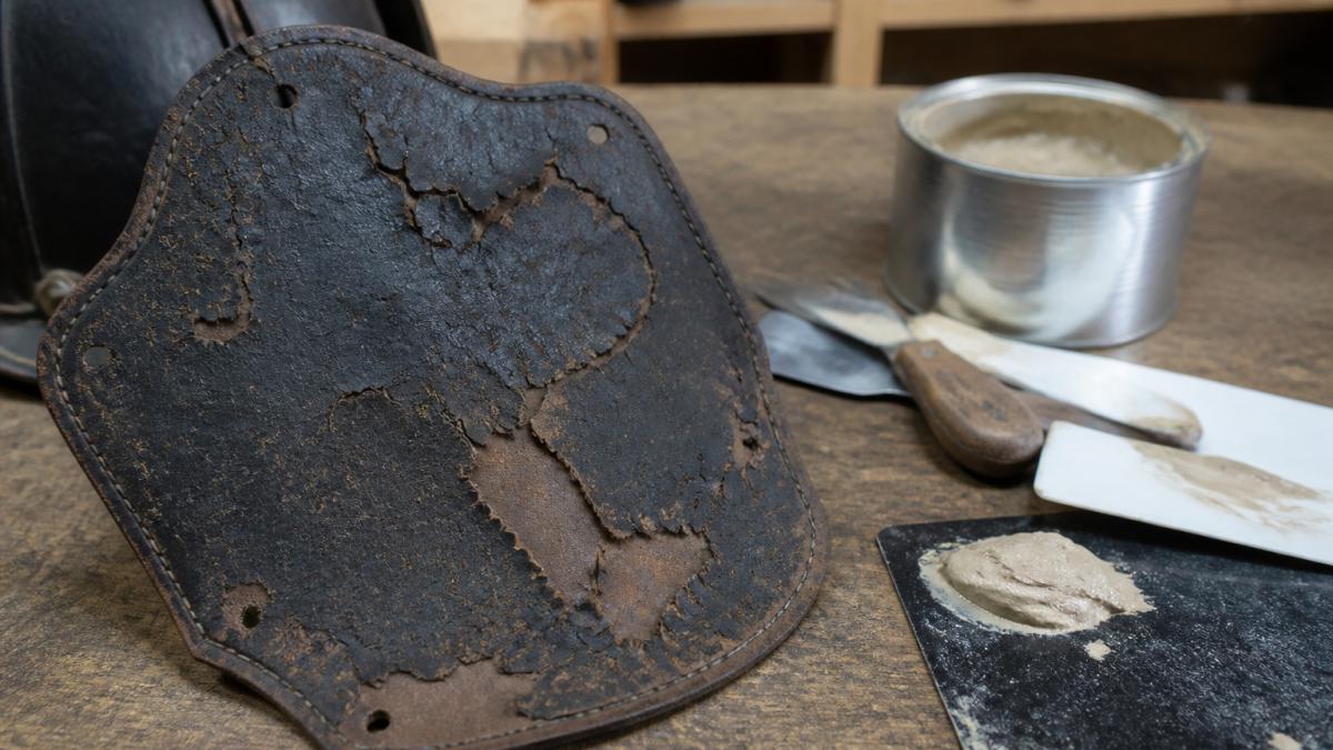 Close-up of a vintage leather fire helmet shield showing visible cracks and areas of delamination with restoration tools and auto-body filler arranged on a work bench nearby