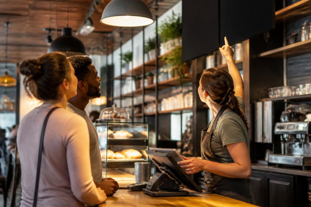Staff member pointing to retail digital screens above a busy café counter as customers choose their orders in a warm setting.