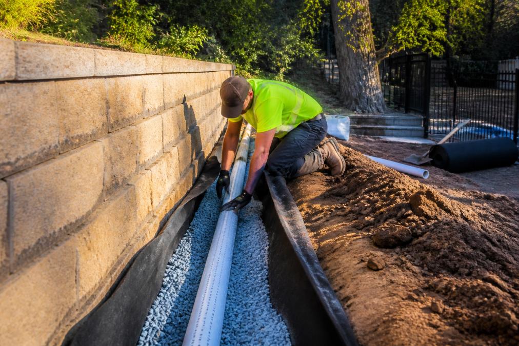 A construction worker in a safety vest kneeling beside a trench behind a retaining wall on a sloped backyard in Sydney, laying a perforated drainage pipe surrounded by blue metal gravel, with geotextile fabric visible and late afternoon sunlight casting long shadows