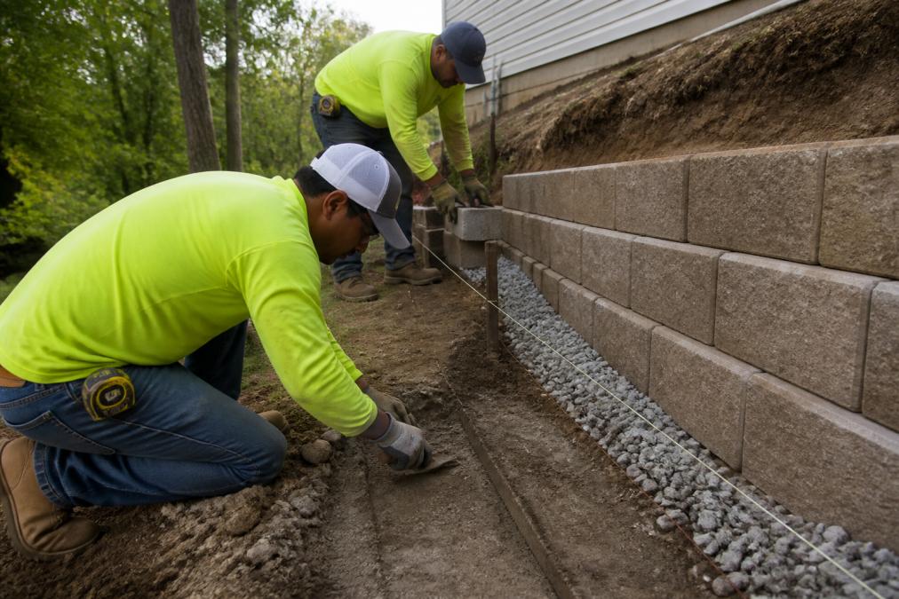 Crew using a mason line during segmental retaining wall installation on a sloped yard with drainage aggregate.