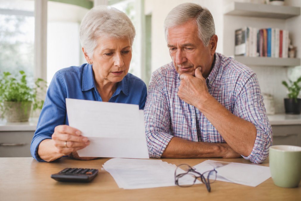 A senior couple sitting at a kitchen table reviewing financial documents with a calculator and reading glasses, natural daylight coming through a window