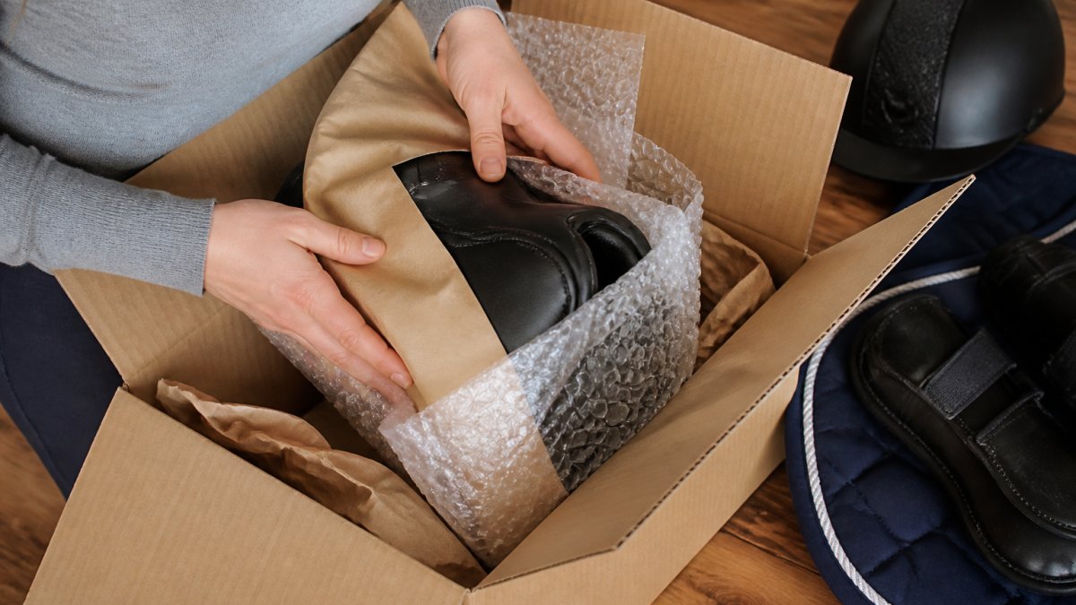 Close-up of hands carefully wrapping equestrian equipment in protective bubble wrap and kraft paper inside a cardboard box, showing proper packaging technique