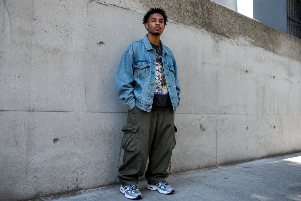 A person wearing an oversized washed denim jacket over a bold graphic t-shirt, paired with baggy cargo pants and retro sneakers, standing against a concrete urban wall in natural daylight