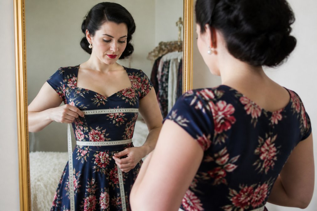 Woman in vintage-inspired dress standing in front of a mirror with a soft measuring tape draped across her bust and waist, showing proper measurement posture and placement