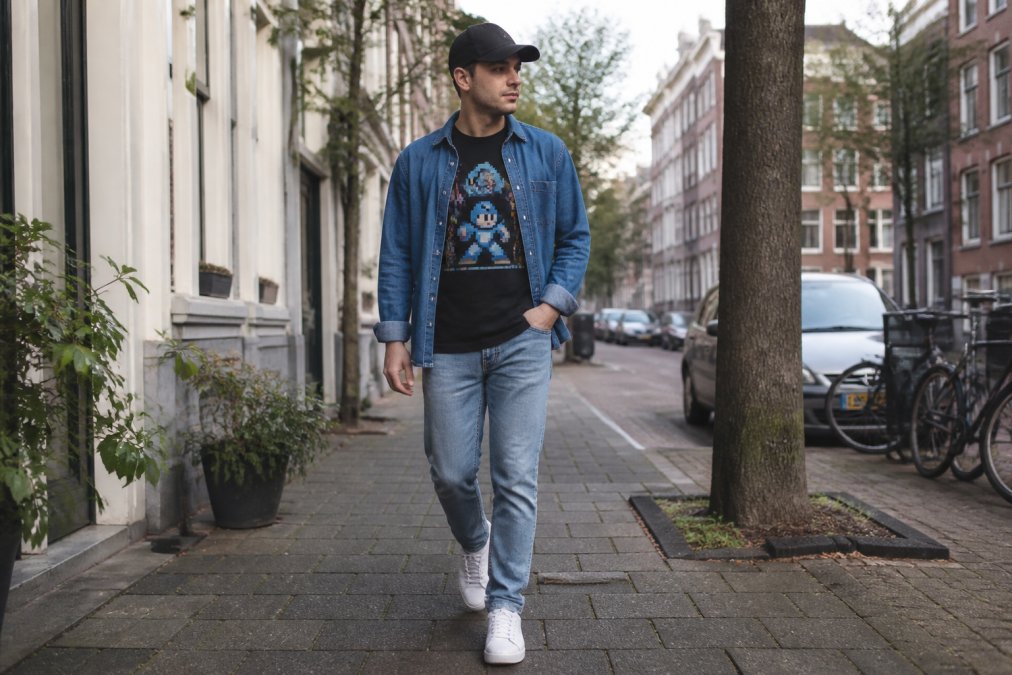 Persoon draagt casual gaming outfit met graphic tee onder open denim overshirt, neutrale jeans, witte sneakers. Moderne, verzorgde look zonder overdreven effect. Natuurlijke daglicht in stadsmilieu
