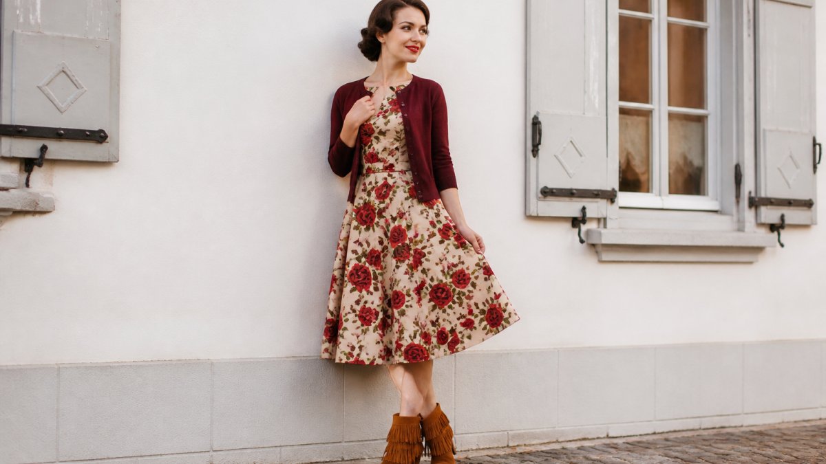 Frau trägt ein Retro-Kleid mit Rosenmuster, bordeauxrote Strickjacke und cognacfarbene Stiefeletten mit Fransen.