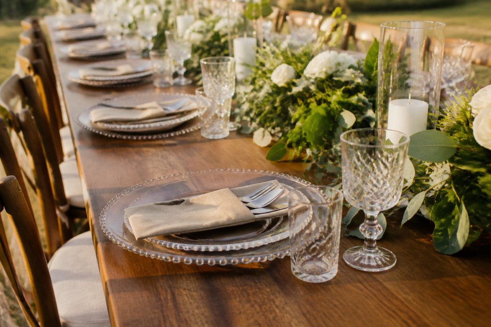 Elegant outdoor wedding reception table with place settings featuring stainless steel utensils in natural pouches, soft golden-hour lighting, green foliage and floral arrangements in background