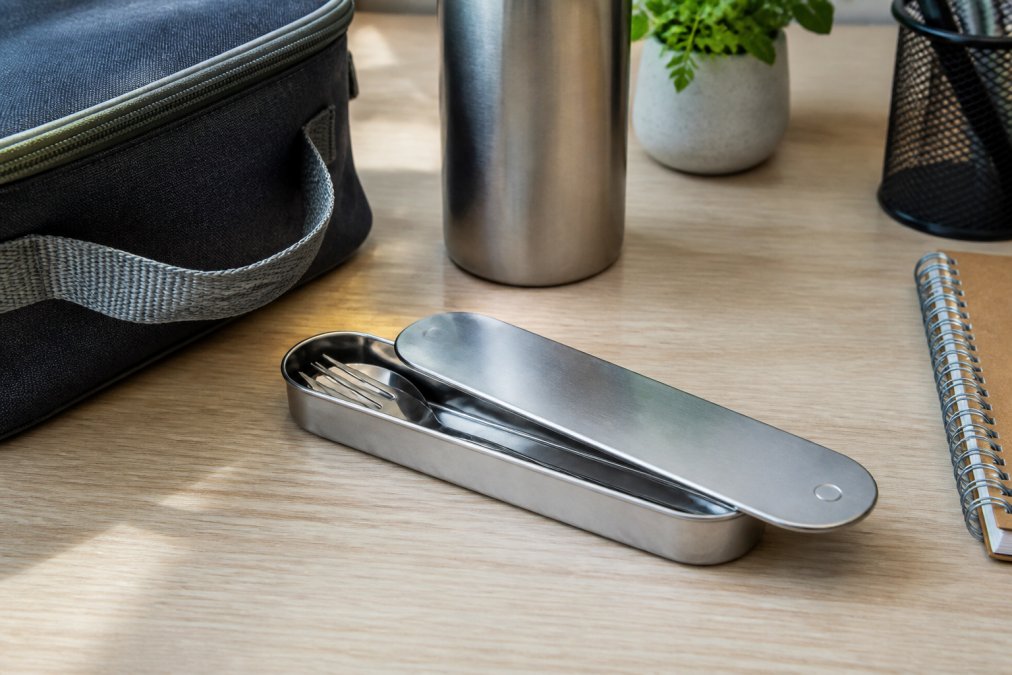 A stainless steel cutlery set with a slider-lid case resting on a desk next to a lunch bag and a reusable water bottle, natural office lighting