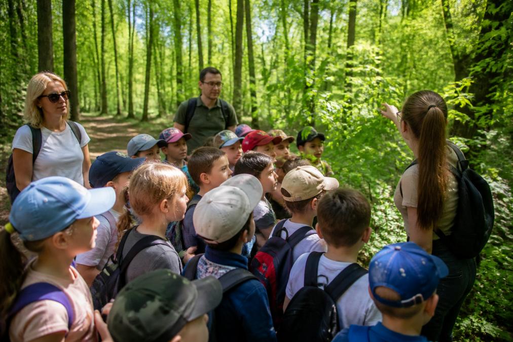 Grupa dzieci na wycieczce szkolnej w parku, pod opieką dorosłych opiekunów, dzieci obserwują naturę i uczestniczą w zajęciach edukacyjnych, słoneczny dzień