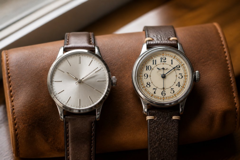Macro photograph of two wristwatches resting side by side on a leather watch roll, one with a clean modern silver dial and one with ornate vintage-style painted numerals, soft window light casting gentle shadows