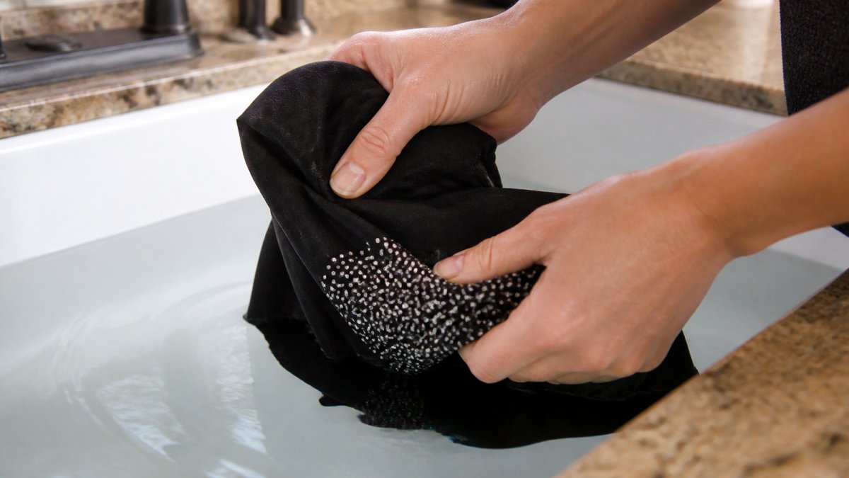 Hands gently turning a rhinestone t-shirt inside out over a sink filled with cool water, showing careful handling of the embellished garment
