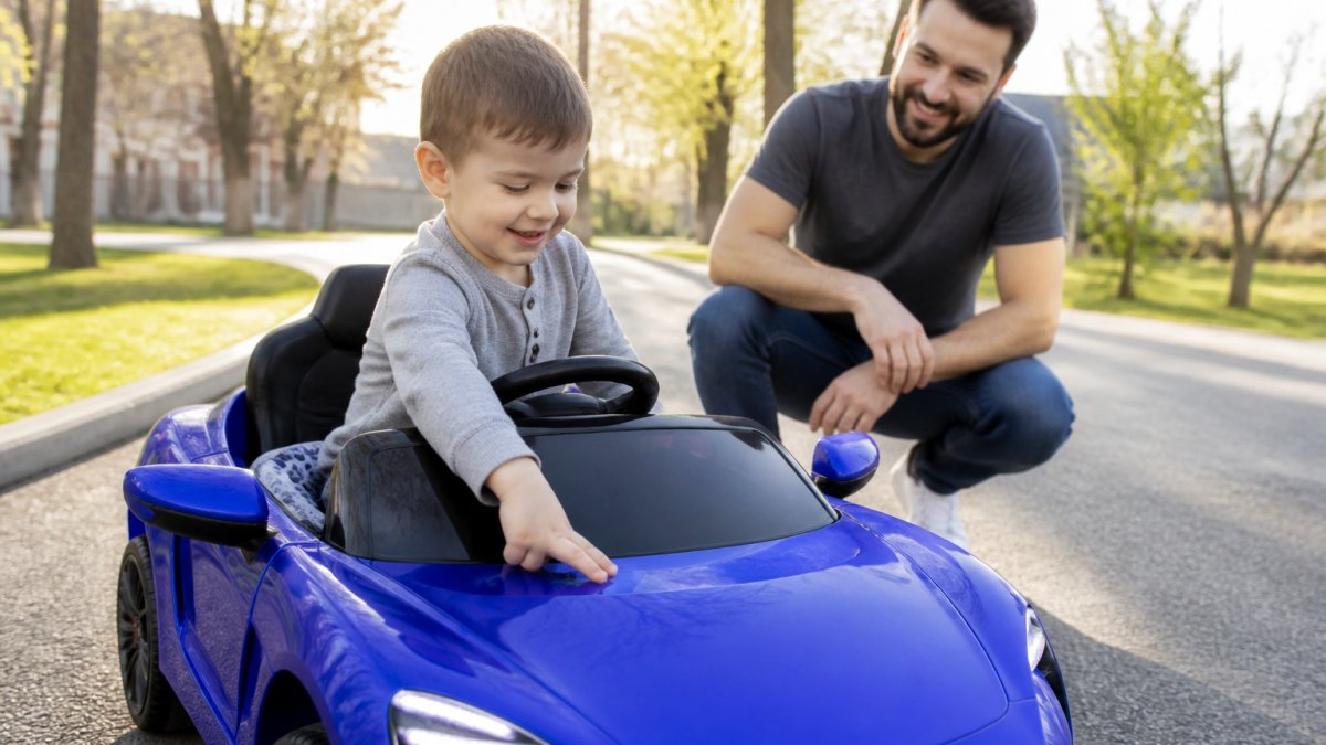 A young child seated in a bright electric ride-on car outdoors, smiling while reaching toward the dashboard controls, with LED lights glowing and a parent supervising nearby in afternoon sunlight