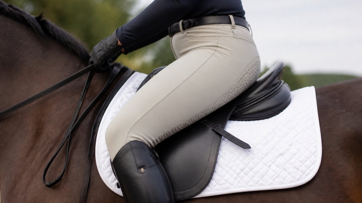 Rider in full-seat riding breeches shown from the side while seated in saddle, demonstrating grip coverage extending from waist to knee with visible silicone texture on inner leg