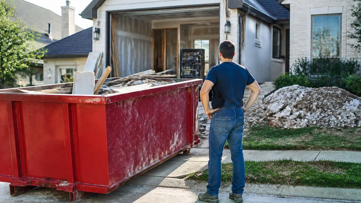 Homeowner standing beside a roll-off dumpster in a residential driveway during an active renovation project, with construction debris visible and the dumpster partially filled, natural daylight