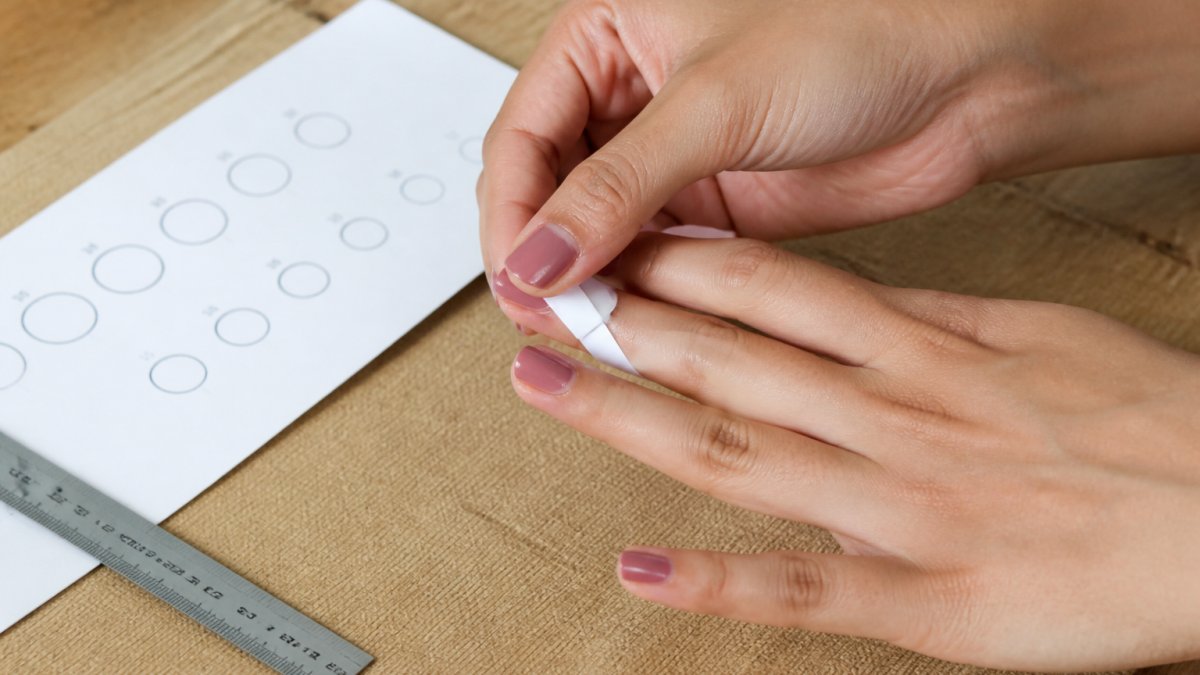 Close-up of hands wrapping a paper strip around a finger to measure ring size, with a printed sizing chart and ruler visible on a wooden surface nearby