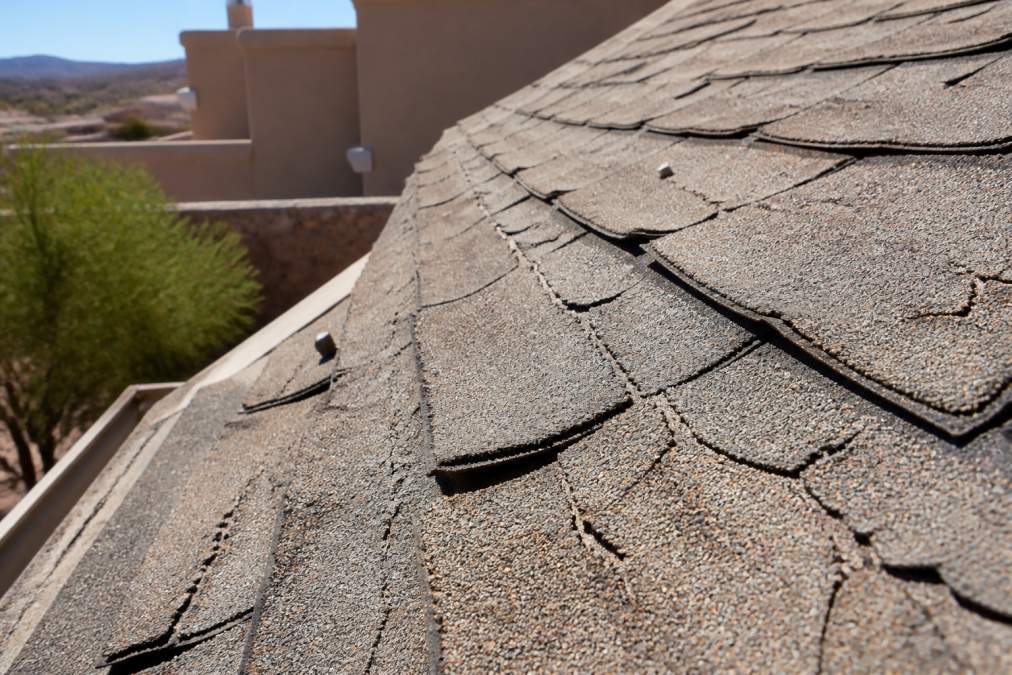Close-up of sun-bleached and cracked asphalt shingles on a residential roof in a dry, sunny desert environment, showing UV damage and brittle granule loss under harsh midday sunlight