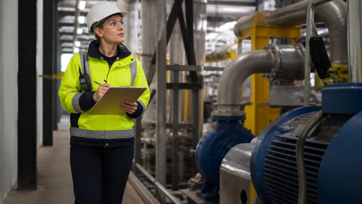 A safety professional in a hard hat and high-visibility vest walking through an industrial plant, clipboard in hand, inspecting equipment and taking notes under overhead lighting