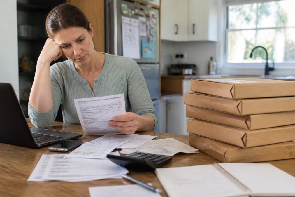 A parent sitting at a kitchen table reviewing a credit card statement on a laptop, with a stack of unopened children's book packages beside them