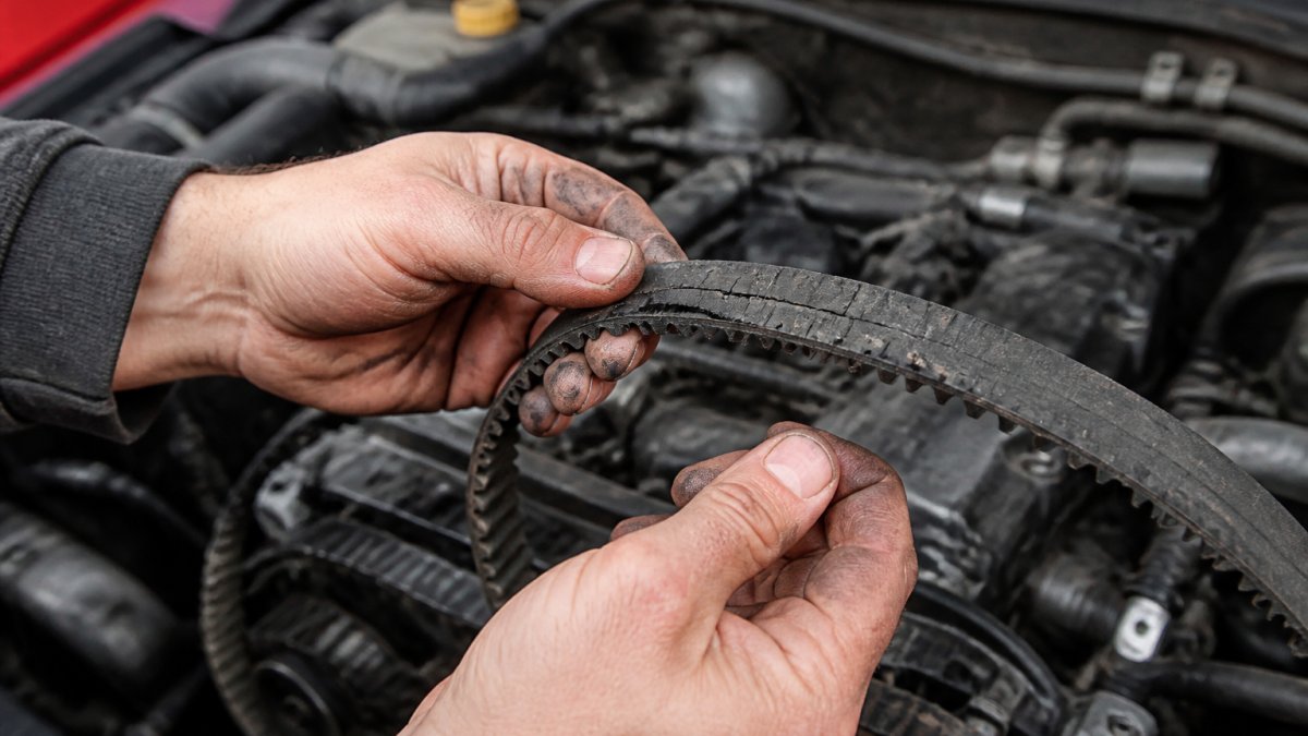 Close-up of a mechanic's hands inspecting a worn timing belt with visible cracks and fraying on the teeth, engine bay in the background, workshop lighting