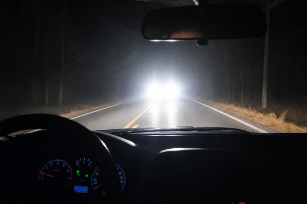 A nighttime scene showing two vehicles facing each other on a dark two-lane road, the oncoming car's LED headlights producing scattered glaring light that washes out the view through the driver's windshield