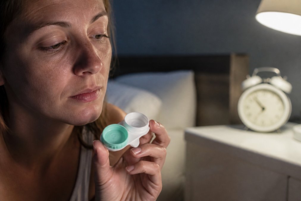 A close-up of a person's hand holding an open contact lens case beside their face, with a bedside clock softly blurred in the background suggesting late evening