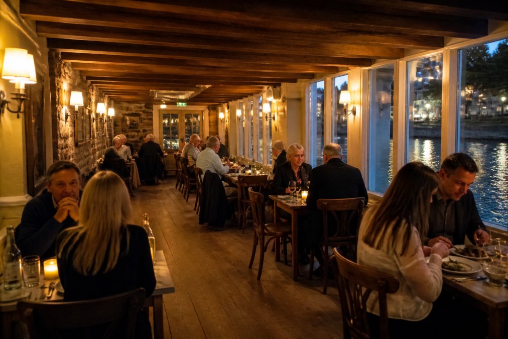 Warm interior of a cosy riverside bistro with wooden beams and historic stone features, diners enjoying meals at candlelit tables with river views visible through tall windows overlooking the waterfront