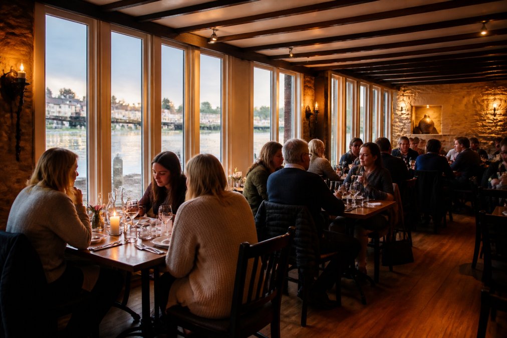 Feature overview showing Warm interior of a cosy riverside bistro with wooden beams and historic stone features, diners en...