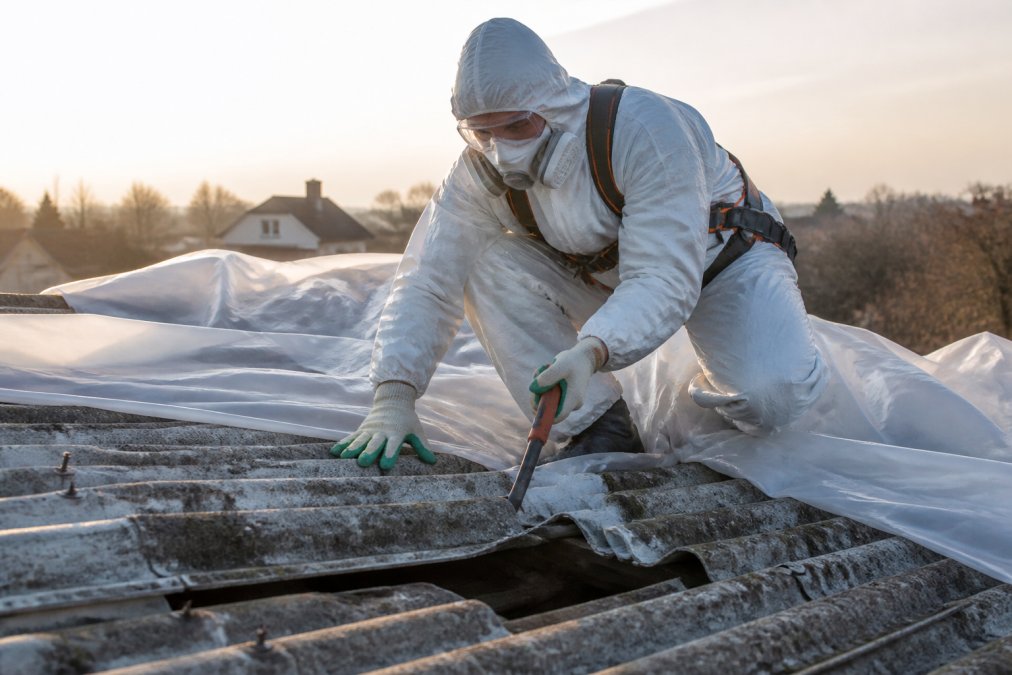 Certifikovaný pracovník v bílém ochranném obleku a respirátoru opatrně demontuje eternitové šablony ze střechy rodinného domu zakryté plachtou, měkké ranní světlo, detail rukavic a nářadí