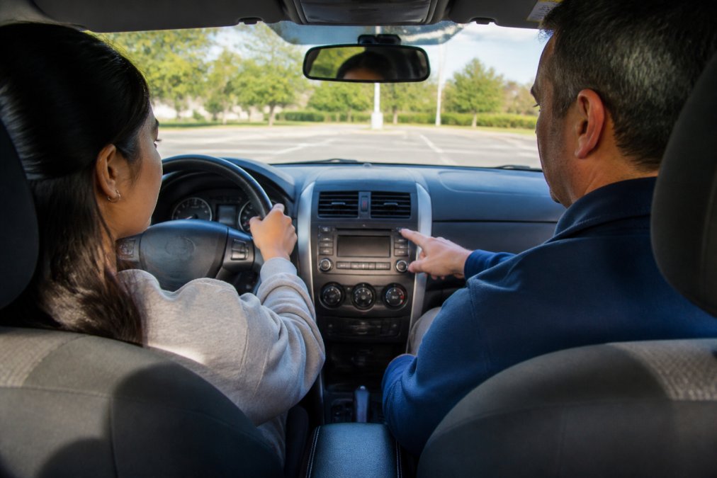 A calm driving instructor sitting in the passenger seat of a sedan, pointing toward the dashboard while a focused international student driver holds the steering wheel, parked in a quiet suburban lot on a clear day