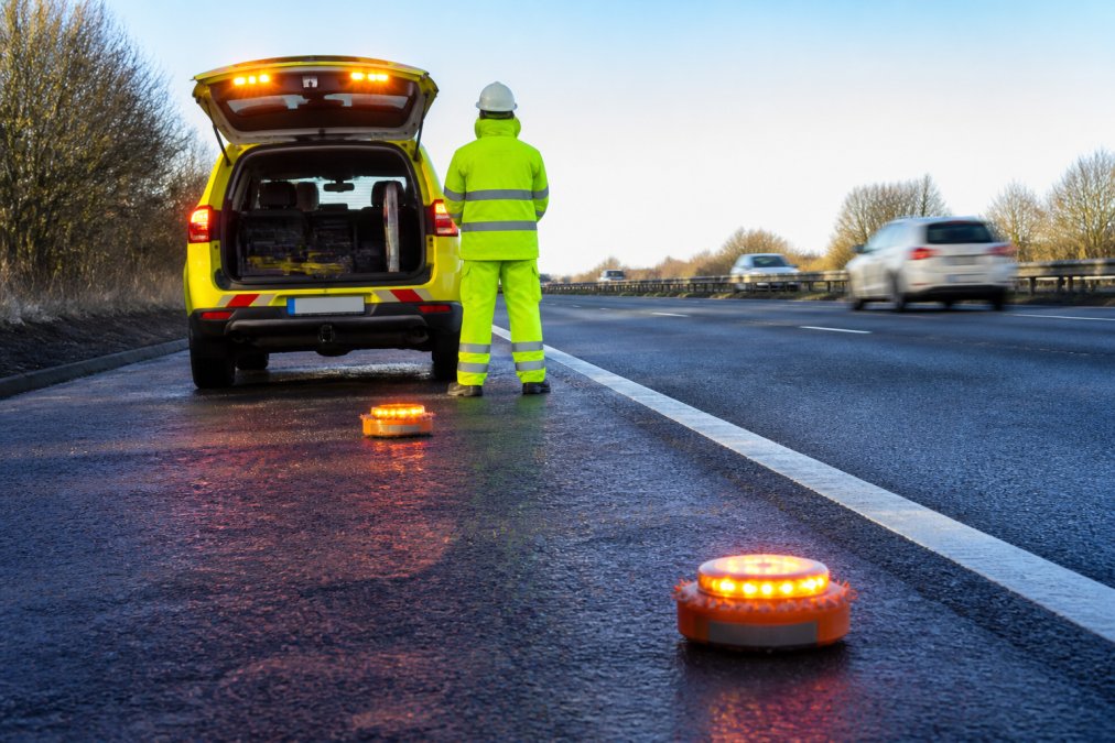 Roadside Safety Devices for Professional Drivers Professional driver in high-visibility clothing standing safely behind a breakdown vehicle on a motorway, with LED warning beacons deployed on the road and hazard lights activated