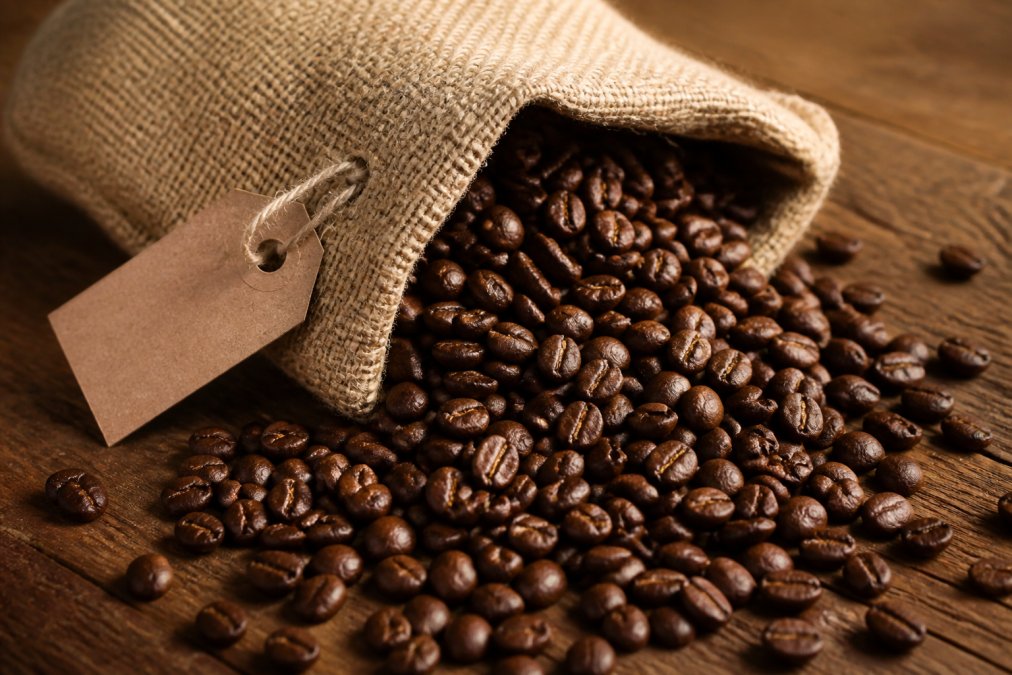 Close-up of a burlap coffee bag with a visible roast date stamp, surrounded by freshly roasted whole beans spilling onto a wooden surface, warm natural lighting