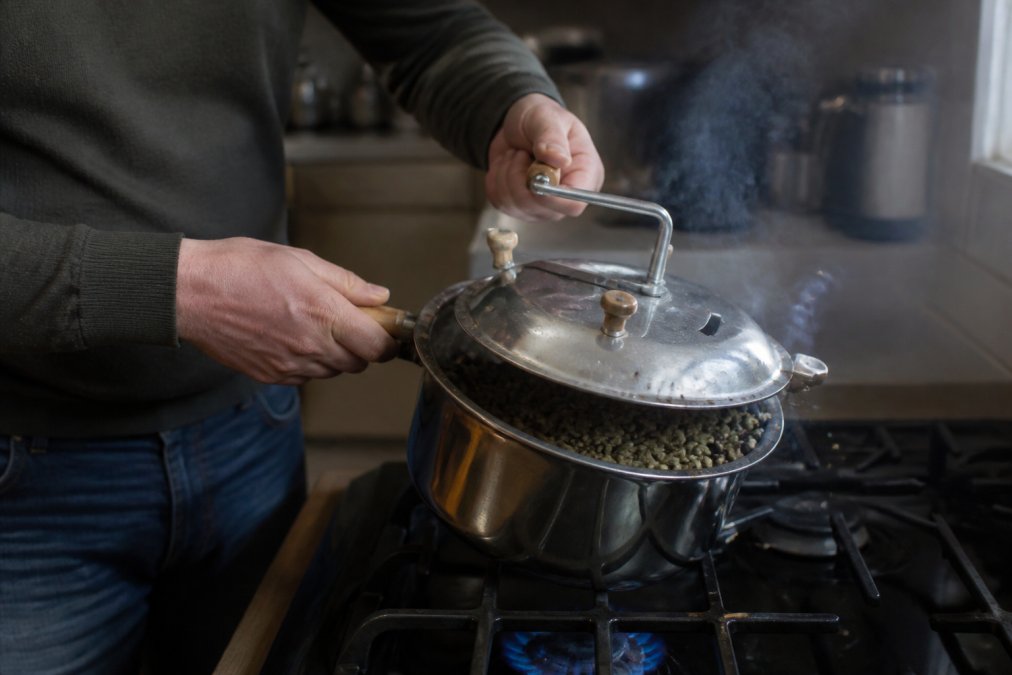 A person using a stovetop popcorn popper to roast green coffee beans, with steam and chaff visible, in a home kitchen setting