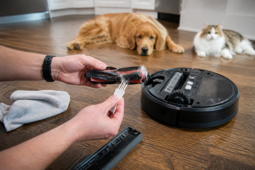 Close-up of a person's hands using a small cleaning tool to remove tangled pet hair from a robot vacuum's roller brush on a kitchen floor, with a dog and cat visible in the background