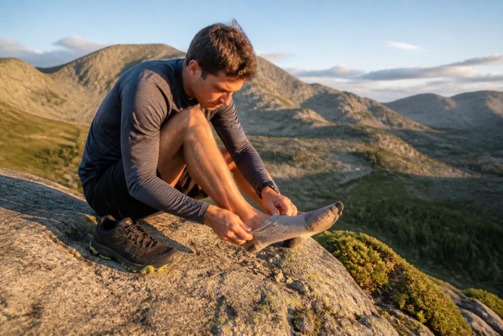 Trail runner sitting on a sun-warmed boulder in a mountain pass, carefully removing their shoe and sock to inspect and remove small pebbles and dust, with a scenic ridge line visible in the soft afternoon light