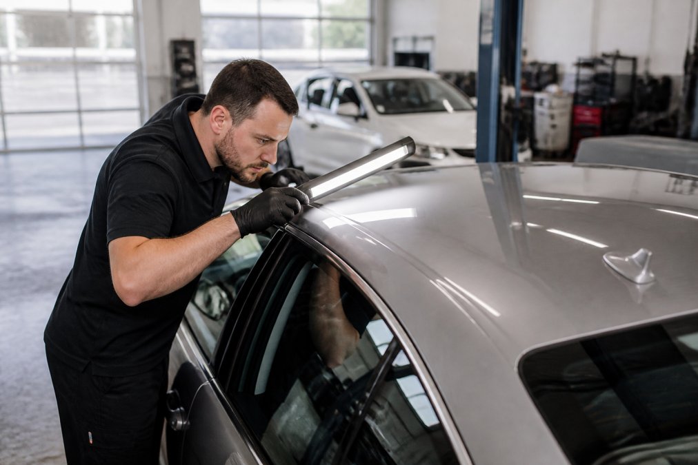 A certified technician in a clean auto body shop inspecting a silver sedan's roof panel with a bright LED light, searching for minor dents under natural shop lighting