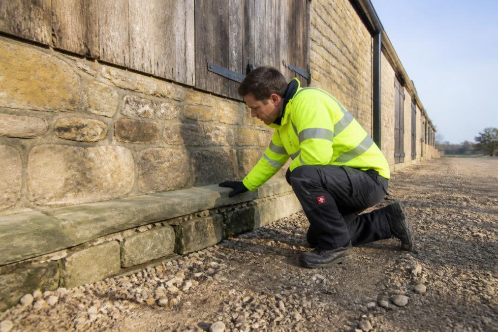 Professional technician in high-visibility clothing inspecting the exterior stone foundation of a large barn, carefully examining gaps and cracks around the base where the stone meets the ground, with morning light illuminating the weathered walls