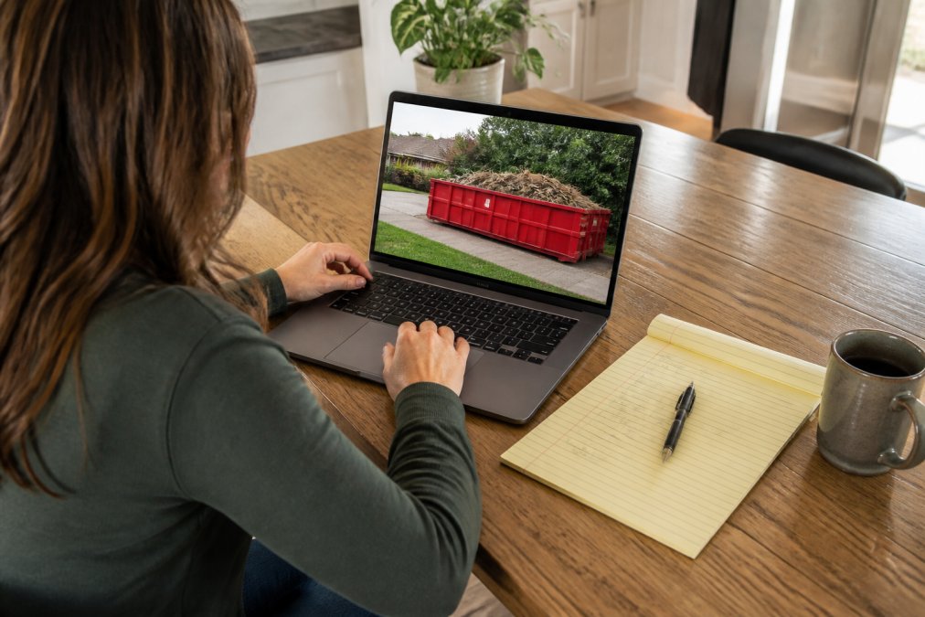 Person on a laptop or mobile device researching and booking a roll-off dumpster rental online, with a notepad showing project details and measurements nearby