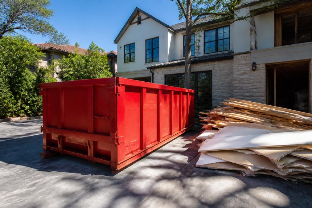 A large red roll-off dumpster parked on a residential driveway next to a two-story home under renovation, with stacked drywall scraps and lumber debris visible beside it, photographed in bright afternoon sunlight