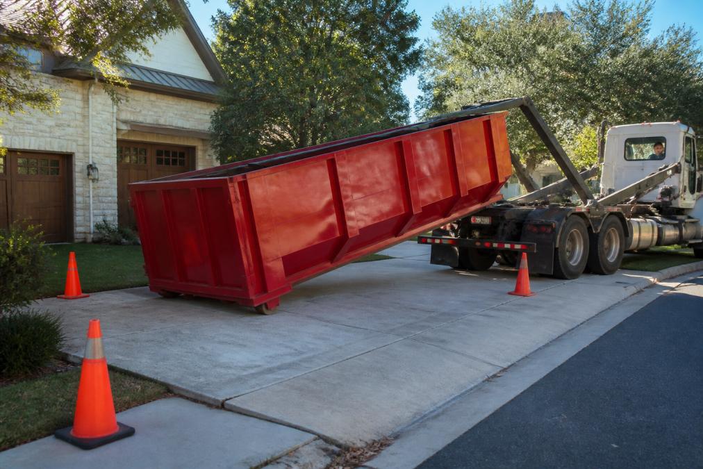 A red roll-off dumpster being lowered from a delivery truck onto a clean, clear residential driveway, with orange traffic cones marking the placement zone and a driver visible in the cab, photographed in morning light