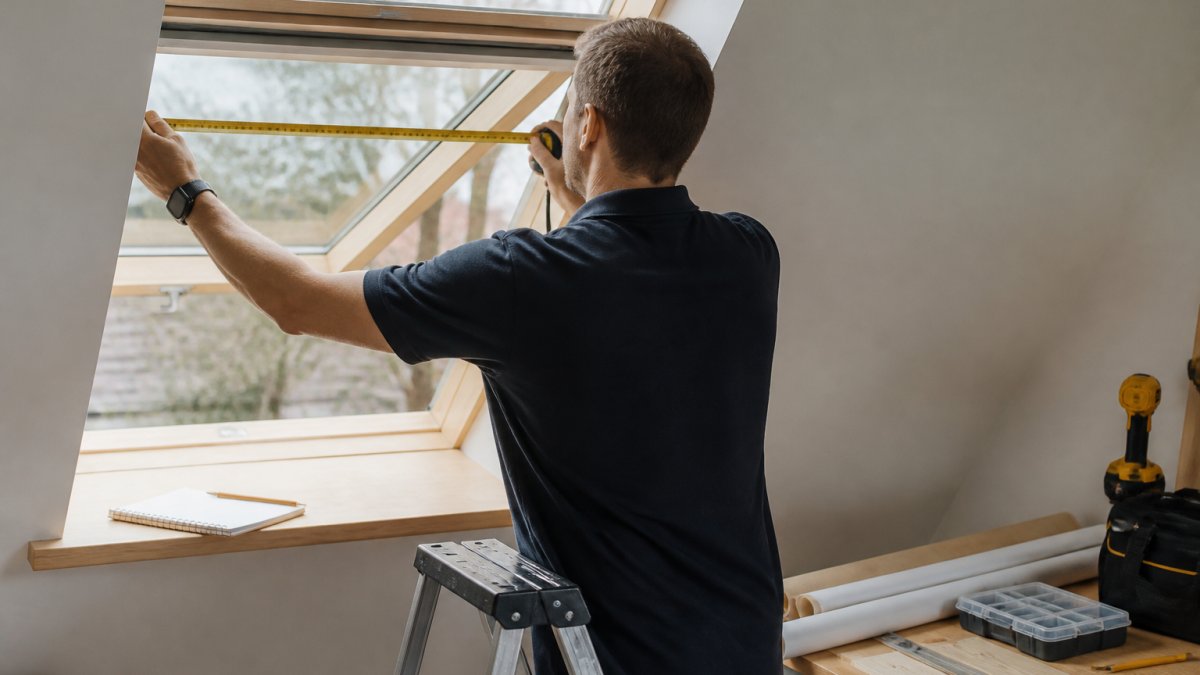 A person standing on a step ladder measuring the width of an open roof window with a metal tape measure, a notebook and pencil resting on the windowsill nearby